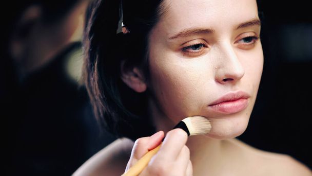 artista de maquillaje borrosa sosteniendo cepillo cosmético mientras se aplica la base de maquillaje en la piel de modelo joven  - Foto, imagen