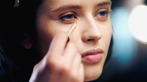 makeup artist holding cosmetic brush while applying concealer on skin of young brunette model  - 写真・画像