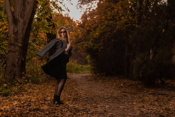 A young girl in a black cloak stands in chameleon glasses in autumn. Yellow forest, green grass, blonde. looking at camera wellbeing concept - Фото, изображение