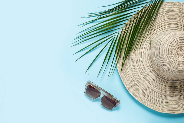 Geniş kenarlı kadın şapkası, güneş gözlüğü ve mavi arka planda bir palmiye dalı. Yaz konsepti, deniz tatili. Pankart. Düz yatış, üst görünüm. - Fotoğraf, Görsel