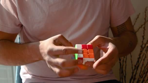 Jovem sentado em uma cadeira no conforto de sua própria casa, resolvendo o quebra-cabeça 3D de um cubo Rubiks. Cara não visível, concentre-se apenas no cubo. Grampo curto - Filmagem, Vídeo