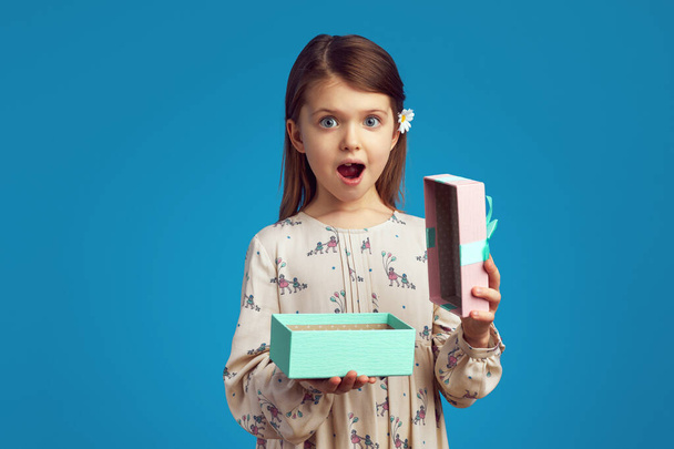 Здивована мила дівчина тримає відкриту подарункову коробку на синьому фоні
 - Фото, зображення