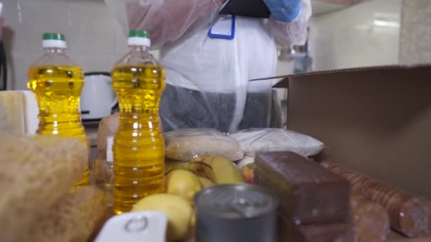 Mercy gönüllüsü, Coronavirus salgını sırasında düşük geliri olan insanlara aktarmak için, koruyucu eldiven takarak, yiyecekleri masadan karton bağış kutusuna koyar. - Video, Çekim