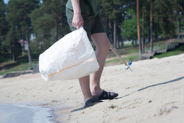 Young guys are engaged in garbage collection on the city beach. Cleaning on the lake in the hot summer. Cleaning is done with a stick with a nail in a white garbage bag. - Foto, Imagem