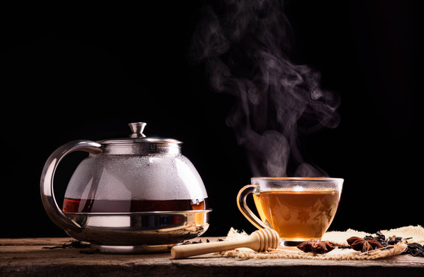 A cup of tea and hot steam from brewed tea with a glass jug with herbs placed on a black background wooden table. - Photo, Image