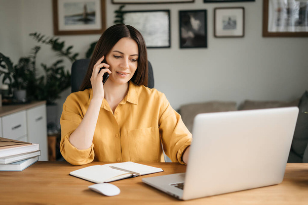 Smiling business woman talking on a mobile phone and using a laptop while working at home, looking at the screen, reading a message, a young female employee advises a client, telephone conversations - Foto, immagini