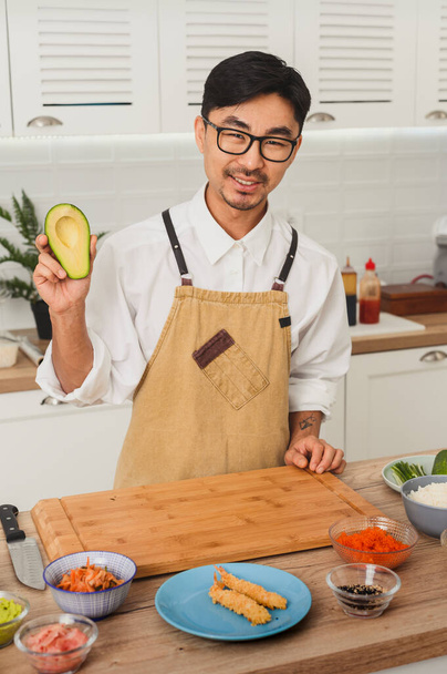 Portret van Aziatische lachende chef-kok in uniform snijden avocado 's voor het maken van sushi - Foto, afbeelding