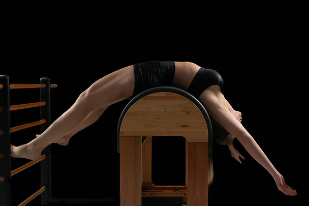Mulher fazendo exercícios pilates em um equipamento de barril, backgound preto, chave baixa. - Foto, Imagem