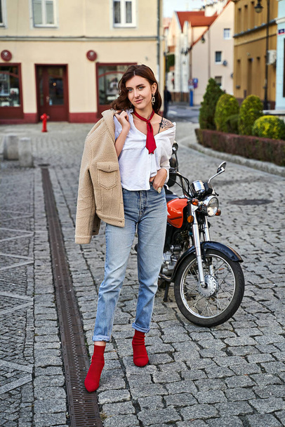 Young beautiful woman in red knitted fabric block heel sock shoes, blue denim jeans pants and teddy jacket coat posing near a motorcycle on a street - Φωτογραφία, εικόνα