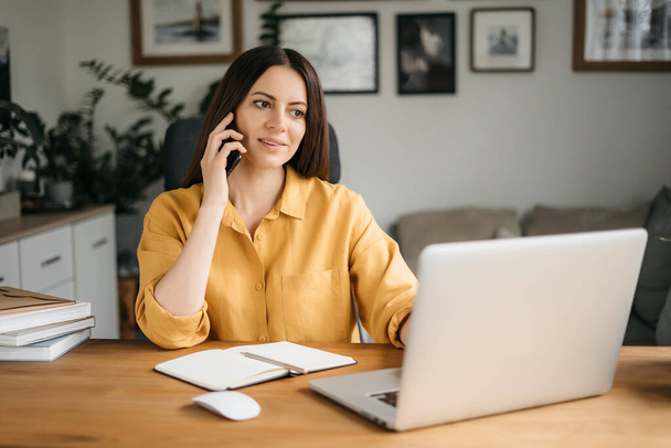 Mujer de negocios sonriente hablando en un teléfono móvil y utilizando un ordenador portátil mientras trabaja en casa, mirando a la pantalla, leyendo un mensaje, una joven empleada aconseja a un cliente, conversaciones telefónicas - Foto, Imagen