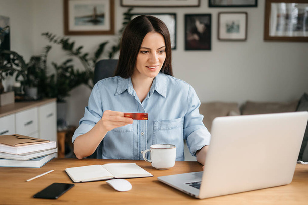 Glücklich lächelnde Frau beim Online-Shopping. Fröhliche Brünette hält Kreditkarte, tätigt Online-Einkäufe, Online-Zahlungen per Laptop, sitzt im Wohnzimmer - Foto, Bild