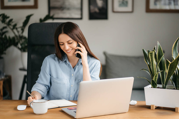 Een glimlachende zakenvrouw die op een mobiele telefoon praat en een laptop gebruikt terwijl ze thuis werkt, naar het scherm kijkt, een bericht leest, een jonge vrouwelijke werknemer adviseert een klant, telefoongesprekken - Foto, afbeelding