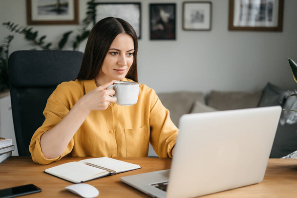 Schöne junge brünette Frau in lässiger gelber Kleidung mit Laptop, während die Arbeit zu Hause Kaffee trinkt - Foto, Bild