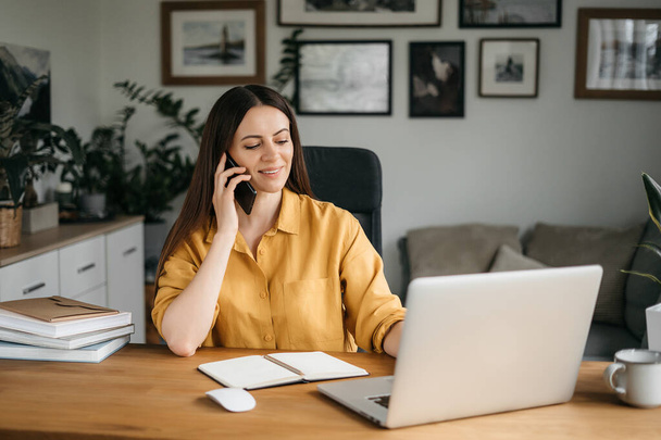 Smiling business woman talking on a mobile phone and using a laptop while working at home, looking at the screen, reading a message, a young female employee advises a client, telephone conversations - Foto, Imagem