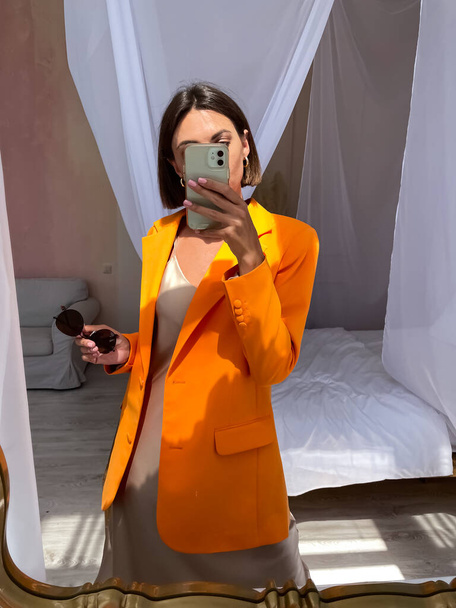 Fit opálená žena v romantických béžových hedvábných šatech a oranžové sako doma fotit selfie na telefonu v zrcadle pro sociální média, příběhy, vertikální - Fotografie, Obrázek