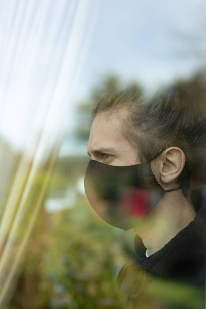 Een jongeman met een gezichtsmasker die thuis uit het raam kijkt. Covid-19 corona virus isolatie eenzaamheid concept - Foto, afbeelding