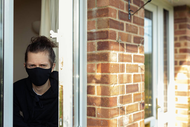 Młody mężczyzna w masce na twarzy, wyglądający przez okno podczas samoizolacji w domu. Koncepcja samotności wirusa Covid-19 - Zdjęcie, obraz