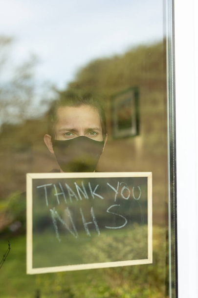 Młody człowiek w masce na twarzy wyglądający przez okno podczas samoizolacji w domu, trzyma znak z podziękowaniami NHS napisanymi na nim - Zdjęcie, obraz