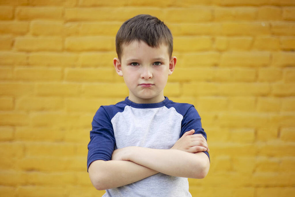 Porträt eines strengen Jungen im T-Shirt, der wütend ist und die Arme vor dem Hintergrund gelber Ziegelwände verschränkt hält. - Foto, Bild