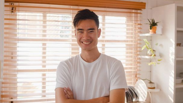 Šťastný mladý Asiat, který se cítí šťastný, usmívá se, ruce zkřížené a dívá se do kamery, zatímco doma odpočívá v kuchyni. Životní styl muž doma koncept. - Fotografie, Obrázek