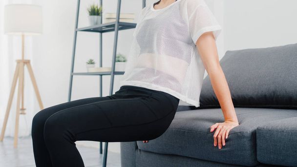 Young Korean lady in sportswear exercises doing working out doing tricep dips leaning on couch in living room at home. Social distance, Isolation during the virus. Exercises for the lower body. - Фото, изображение