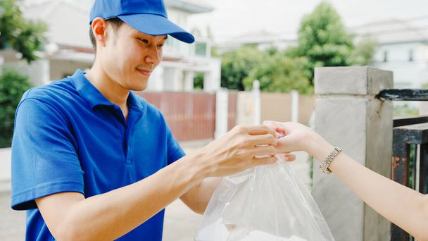 Young Asia postal delivery courier man in blue shirt handling food boxes for sending to customer at house and Asian female receive delivered package outdoors. Package shopping food delivery concept. - Photo, Image