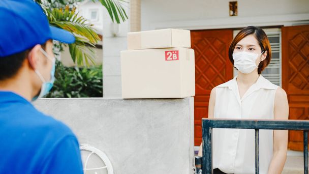 Young postal delivery courier man wear face mask handling parcel box for send to customer at house and Asian female receive delivered package outdoors. Lifestyle new normal after corona virus concept. - Foto, immagini