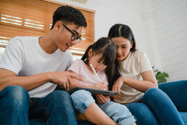 Szczęśliwy azjatycki ojciec rodziny, mama i córka za pomocą komputera tablet technologii siedzi sofa w salonie w domu. Samoizolacja, pozostawanie w domu, dystans społeczny, kwarantanna w celu zapobiegania koronawirusom. - Zdjęcie, obraz