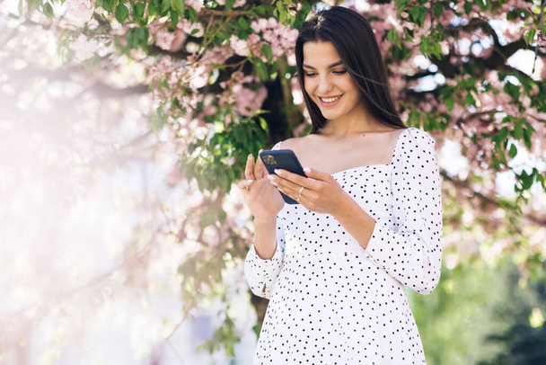 Smiling woman wearing white dress using smartphone. Girl using social media application text messages receive news smiling outdoor. Communication, social networks, online shopping concept. Technology - Fotoğraf, Görsel