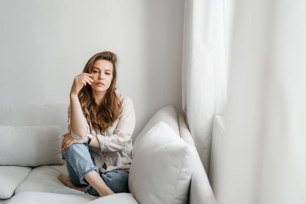 Una joven rubia con ropa cómoda casual se sienta en un sofá blanco en casa. interior escandinavo, minimalismo - Foto, Imagen