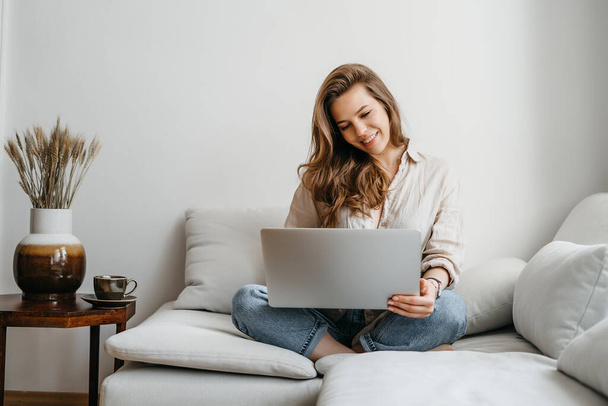 Portrét mladé ženy na volné noze sedící na gauči a šťastně používající notebook doma - Fotografie, Obrázek