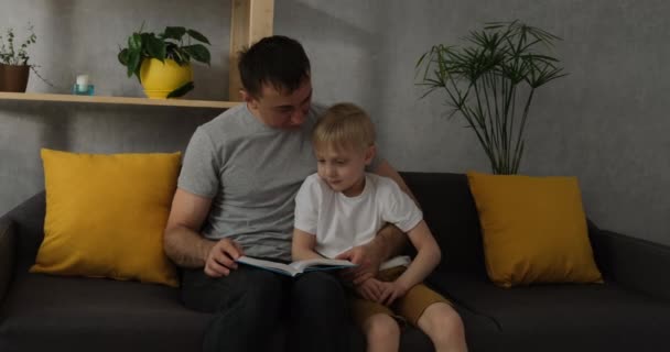Otec čte knihu svému synovi, chlapec pozorně poslouchá. Táta tráví čas s dítětem. Školka s tátou si doma na gauči čtou. - Záběry, video