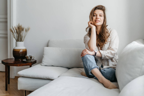 Una joven rubia con ropa cómoda y casual se sienta en un sofá blanco en casa - Foto, Imagen
