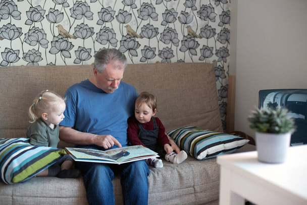 Ανώτερος άντρας διαβάζει βιβλία σε μικρά κορίτσια. Ο παππούς περνάει χρόνο με εγγονές στον καναπέ.. - Φωτογραφία, εικόνα