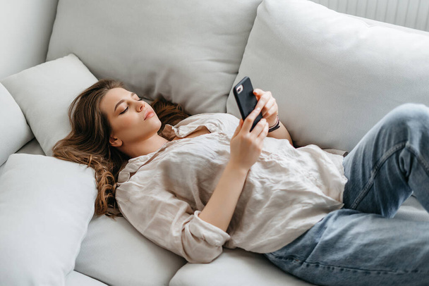 Νεαρή γυναίκα κοιτάζει στην οθόνη του τηλεφώνου, ενώ βρίσκεται στον καναπέ στο σπίτι - Φωτογραφία, εικόνα