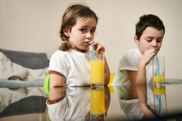 Cute children drinking multi vitamin juice from a straw sitting at the table in the living room - 写真・画像
