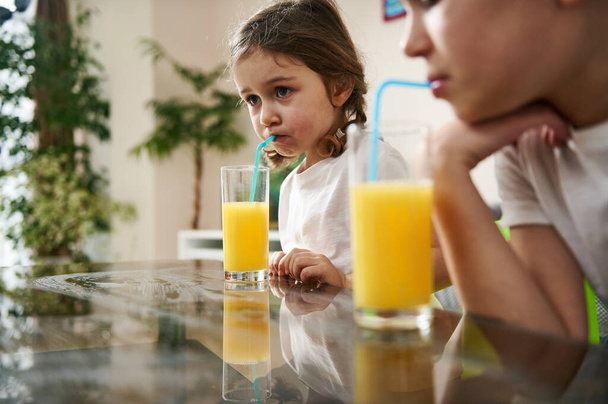 Adorable baby girl drinking orange juice sitting at the table at home next to her brother blurred in the foreground - Foto, afbeelding