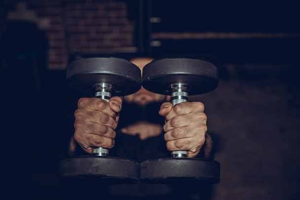 Close up of a muscular young man lifting weights in gym on dark background. - Photo, Image