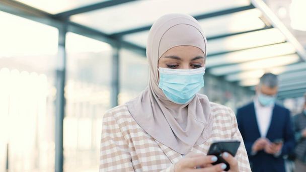 Close up of Muslim pretty concentrated woman in medical mask standing on street outdoors at bus stop and texting on smartphone gadget browsing online. Female typing on mobile phone in city - Foto, immagini