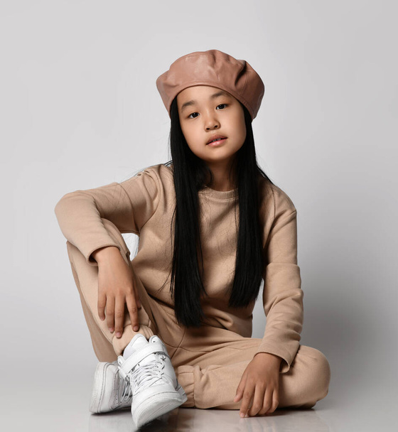 Asian brunette kid girl in green hat beret, pants, hoodie, sweater and sneakers sits on floor looking at camera - Fotó, kép