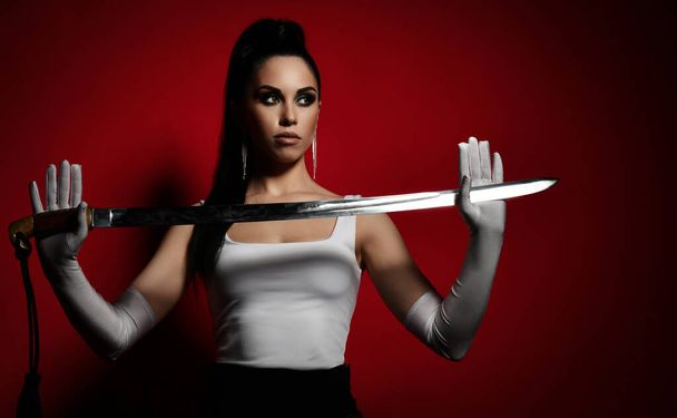 Young pretty brunette woman in white top and gloves standing and holding dagger in hands looking away - Foto, imagen