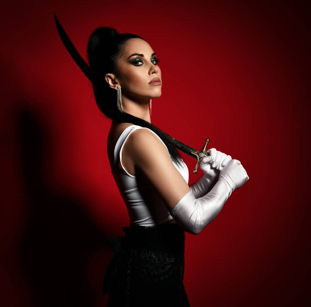 Portrait of young pretty brunette serious female in white gloves standing and holding asian dagger in hands - Foto, Imagem
