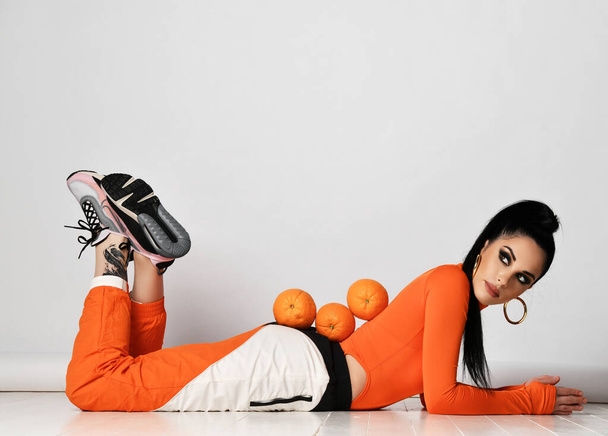 Beautiful positive young brunette woman in bright orange sport costume and smeakers lying on floor with fresh oranges on back - 写真・画像