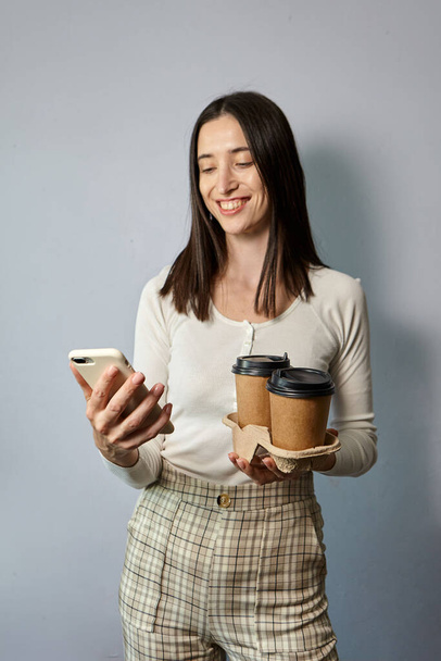 Eine Frau im weißen Sweatshirt hält in der einen Hand zwei Tassen Kaffee, in der anderen ein Smartphone. Tee oder Kaffee zum Mitnehmen. Fernkommunikation - Foto, Bild