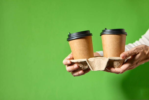 Χέρια κρατώντας δύο φλιτζάνια καφέ χαρτί με μαύρο καπάκι. Δύο ειδικές προσφορές καφέ ή promo. Χέρια κρατώντας δύο φλιτζάνια σε πράσινο φόντο. Τσάι ή καφέ για έξω. Καφέ χάρτινο κύπελλο με μαύρο καπάκι. - Φωτογραφία, εικόνα
