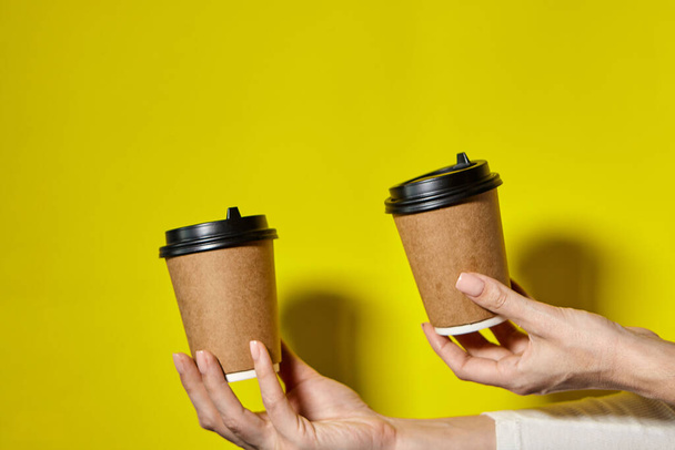 Hands holding two cups brown paper with black lid. Two coffee special offer or promo. Hands holding two cups on yellow screen background. Tea or Coffee to go. Brown paper cup with black lid. - Foto, Imagem
