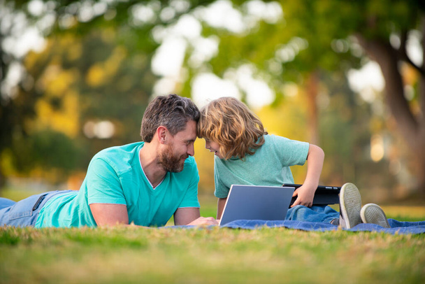 Happy father with son use laptop. Dad and school boy child looking screen and tablet, watching video lesson, sitting on grass. Family weekend. Video call of a grandson to grandparents. - Foto, Bild
