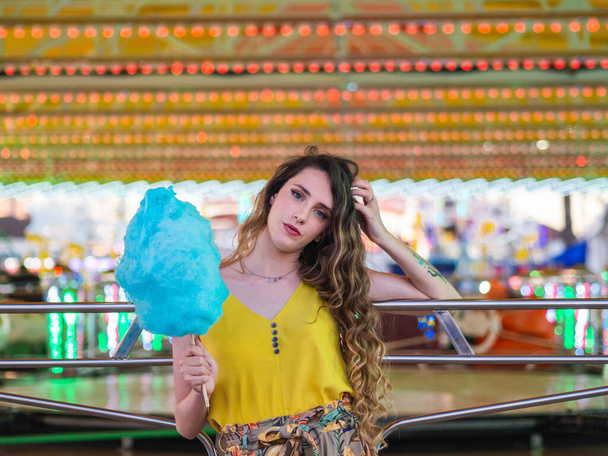 A beautiful caucasian girl posing with blue cotton candy in front of the bumping cars at a park - Photo, image