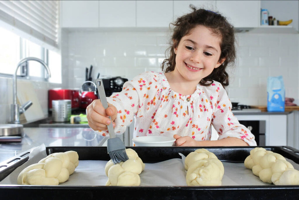 Happy young Jewish girl (female age 7) baking sweet Challah bread for Sabbath Jewish Holiday in home kitchen. - Foto, Bild