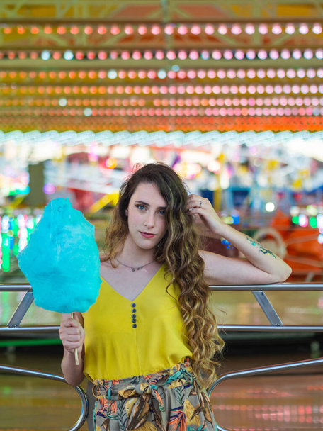 A beautiful caucasian girl posing with blue cotton candy in front of the bumping cars at a park - Φωτογραφία, εικόνα